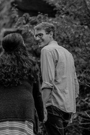 A black and white photograph of a couple holding hands, walking outdoors in a natural setting. The man, with short hair and glasses, is looking back at the woman with a smile. The woman has long curly hair and is wearing a striped skirt and dark top. The background has blurred foliage, giving a sense of being in a forest or garden.