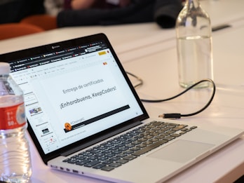 A laptop displaying a presentation slide with Spanish text about certificate distribution. Beside the laptop, there is a plastic bottle partially filled with water and a glass bottle. The setting appears to be a meeting or workshop environment.