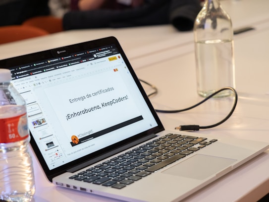 A laptop displaying a presentation slide with Spanish text about certificate distribution. Beside the laptop, there is a plastic bottle partially filled with water and a glass bottle. The setting appears to be a meeting or workshop environment.