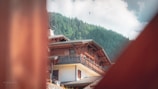 A view of a wooden chalet-style building with a forested hillside in the background. There are several paragliders visible in the sky, providing a sense of outdoor adventure and leisure. The image is framed by blurred wooden structures in the foreground, adding depth to the composition.
