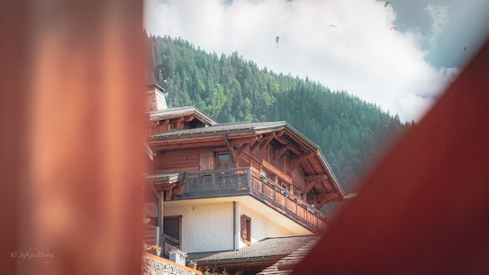 A view of a wooden chalet-style building with a forested hillside in the background. There are several paragliders visible in the sky, providing a sense of outdoor adventure and leisure. The image is framed by blurred wooden structures in the foreground, adding depth to the composition.