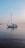 A small sailboat is gently moving across a calm body of water during what appears to be either sunrise or sunset. The sky is a gradient of soft blues and pinks, reflecting peacefully on the water's surface. A couple of people can be seen on the boat, involved in operating it.