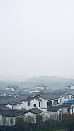 A serene landscape featuring traditional Chinese architecture with white walls and dark-tiled roofs. The houses are nestled amidst greenery, with trees and plants surrounding the area. A misty sky creates a tranquil atmosphere, enhancing the peacefulness of the scene.
