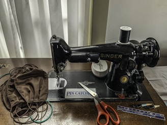 A vintage sewing machine is placed on a wooden table. Adjacent to the machine are several brown fabric pieces, a pair of orange scissors, a measuring tape, and some sewing accessories. The machine brand 'Singer' is visible and it is set against a backdrop of grey curtains.