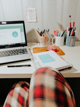 A workspace with a laptop displaying a website, surrounded by stationary items including pens and markers in mugs. Notebooks are open with a post-it note on top, and there is a person's knee in plaid pajama pants in the foreground.