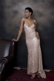 A person in a sparkling, sleeveless gown poses elegantly against a dark background. The dress is long and form-fitting with shimmering details, and the person leans slightly on a dark brown, leather armchair. The setting is subdued and stylish, providing a contrast that highlights the glittering gown.
