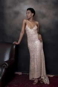 A person in a sparkling, sleeveless gown poses elegantly against a dark background. The dress is long and form-fitting with shimmering details, and the person leans slightly on a dark brown, leather armchair. The setting is subdued and stylish, providing a contrast that highlights the glittering gown.