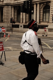 A person dressed in a uniform with a red cap, white shirt, and dark pants is walking on a city street. The individual appears to be wearing a mask and carrying a bag. The background features a building with large, arched windows and traffic lights. Street signs and traffic cones are visible, indicating some construction or road blockages.