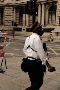 A person dressed in a uniform with a red cap, white shirt, and dark pants is walking on a city street. The individual appears to be wearing a mask and carrying a bag. The background features a building with large, arched windows and traffic lights. Street signs and traffic cones are visible, indicating some construction or road blockages.