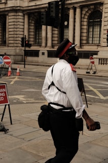 A person dressed in a uniform with a red cap, white shirt, and dark pants is walking on a city street. The individual appears to be wearing a mask and carrying a bag. The background features a building with large, arched windows and traffic lights. Street signs and traffic cones are visible, indicating some construction or road blockages.