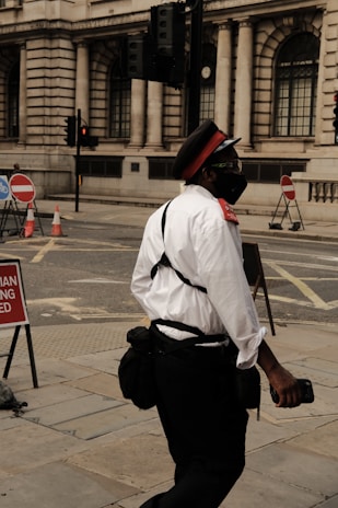 A person dressed in a uniform with a red cap, white shirt, and dark pants is walking on a city street. The individual appears to be wearing a mask and carrying a bag. The background features a building with large, arched windows and traffic lights. Street signs and traffic cones are visible, indicating some construction or road blockages.