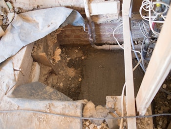 A construction or renovation site with a partially excavated area surrounded by wooden beams and metal pipes. Exposed wiring is visible to the right, and a brick wall forms the backdrop. Dust and debris are scattered around the site, with a partially removed tarp draped over one side.