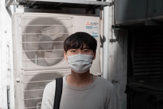 A person wearing a face mask stands in front of an industrial air conditioning unit. The background includes metallic structures and pipes, suggesting an outdoor urban setting. The individual is dressed in a light sweater and carries a bag over one shoulder.