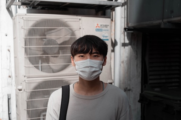A person wearing a face mask stands in front of an industrial air conditioning unit. The background includes metallic structures and pipes, suggesting an outdoor urban setting. The individual is dressed in a light sweater and carries a bag over one shoulder.