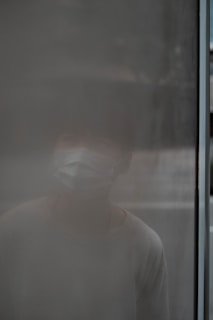A person wearing a mask is barely visible behind a foggy or frosted glass surface, creating an obscure and mysterious effect.