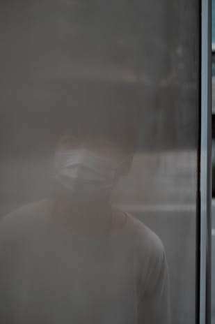 A person wearing a mask is barely visible behind a foggy or frosted glass surface, creating an obscure and mysterious effect.