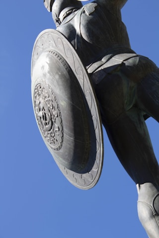 A large statue features a warrior figure with intricate armor and a detailed round shield, set against a clear blue sky. The perspective is slightly angled upwards, highlighting the grandeur and ornate design of the shield and the dynamic posture of the figure.