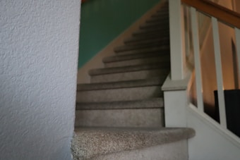 A carpeted staircase with a wooden handrail and white balusters leads upward, flanked by textured walls. The lighting is soft and casts subtle shadows on the stairs and the wall.
