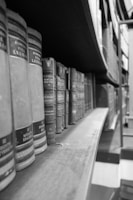 Rows of classic, hardcover books are neatly aligned on a wooden shelf in a monochromatic setting, creating a vintage library atmosphere.