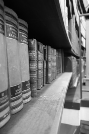 Rows of classic, hardcover books are neatly aligned on a wooden shelf in a monochromatic setting, creating a vintage library atmosphere.