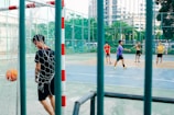 A group of young men are playing futsal on an outdoor court. One player in a black outfit, near the net, appears to have scored a goal as the orange ball is in the goal. The other players are spread across the court, with one wearing a red shirt and another in a purple shirt, participating actively in the game. The court is enclosed by a high fence and surrounded by greenery and buildings.