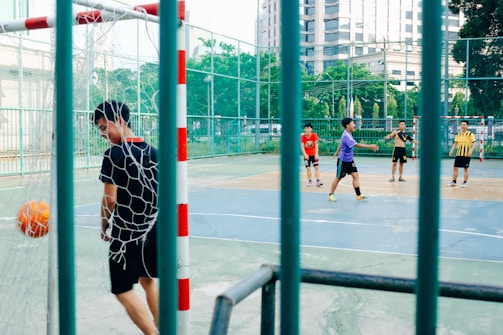 A group of young men are playing futsal on an outdoor court. One player in a black outfit, near the net, appears to have scored a goal as the orange ball is in the goal. The other players are spread across the court, with one wearing a red shirt and another in a purple shirt, participating actively in the game. The court is enclosed by a high fence and surrounded by greenery and buildings.