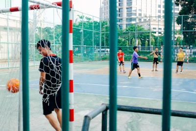 A group of young men are playing futsal on an outdoor court. One player in a black outfit, near the net, appears to have scored a goal as the orange ball is in the goal. The other players are spread across the court, with one wearing a red shirt and another in a purple shirt, participating actively in the game. The court is enclosed by a high fence and surrounded by greenery and buildings.