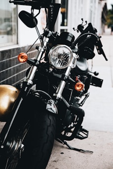 A close-up view of a black motorcycle parked on a sidewalk, showcasing its front headlight and handlebars. The motorcycle features chrome accents and orange turn signals, with a reflective surface visible on the body and a background of storefronts and buildings.