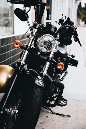A close-up view of a black motorcycle parked on a sidewalk, showcasing its front headlight and handlebars. The motorcycle features chrome accents and orange turn signals, with a reflective surface visible on the body and a background of storefronts and buildings.