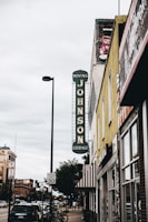 A street view featuring a vintage vertical sign that reads 'Johnson Storage Moving' on the side of a building. The surrounding area includes various storefronts, parked cars along the sidewalk, and a speed limit sign indicating 30 mph. A painted teddy bear mural can be seen near the top of one building, and the sky is overcast, adding a muted tone to the scene.