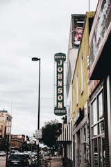A street view featuring a vintage vertical sign that reads 'Johnson Storage Moving' on the side of a building. The surrounding area includes various storefronts, parked cars along the sidewalk, and a speed limit sign indicating 30 mph. A painted teddy bear mural can be seen near the top of one building, and the sky is overcast, adding a muted tone to the scene.