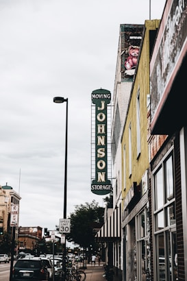 A street view featuring a vintage vertical sign that reads 'Johnson Storage Moving' on the side of a building. The surrounding area includes various storefronts, parked cars along the sidewalk, and a speed limit sign indicating 30 mph. A painted teddy bear mural can be seen near the top of one building, and the sky is overcast, adding a muted tone to the scene.