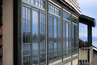 A series of large rectangular windows with a grid-like pattern are prominently displayed on the side of a building. The window frames are green and reflect an outdoor scene, including a blurred view of a lake or sea and some trees. The building has a shingled roof and is situated in a serene, natural setting.