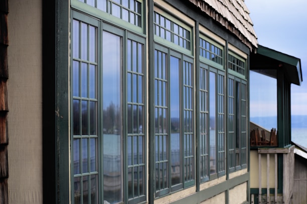 A series of large rectangular windows with a grid-like pattern are prominently displayed on the side of a building. The window frames are green and reflect an outdoor scene, including a blurred view of a lake or sea and some trees. The building has a shingled roof and is situated in a serene, natural setting.