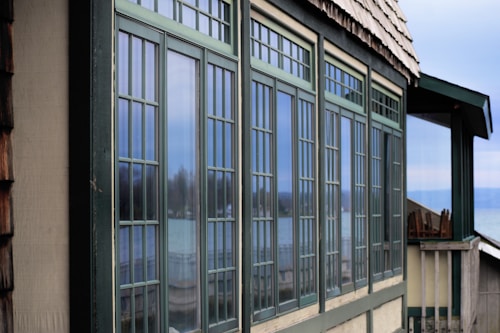 A series of large rectangular windows with a grid-like pattern are prominently displayed on the side of a building. The window frames are green and reflect an outdoor scene, including a blurred view of a lake or sea and some trees. The building has a shingled roof and is situated in a serene, natural setting.