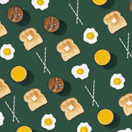 A patterned arrangement of breakfast items including slices of toast with butter, fried eggs, glasses of orange juice, iced coffee, and striped straws spread across a dark green background.