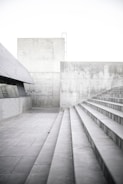 Concrete steps lead upward alongside a gray, modern architectural structure. The walls and floors are made of concrete, creating a minimalist and industrial setting. The lighting is bright and soft, highlighting the texture of the concrete surfaces.