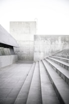 Concrete steps lead upward alongside a gray, modern architectural structure. The walls and floors are made of concrete, creating a minimalist and industrial setting. The lighting is bright and soft, highlighting the texture of the concrete surfaces.