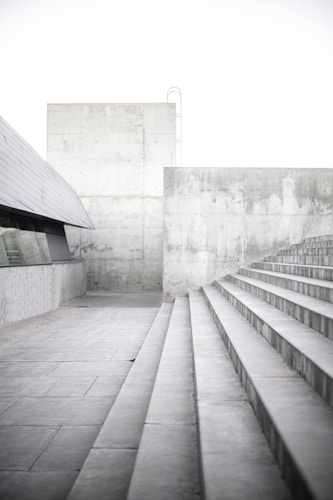 Concrete steps lead upward alongside a gray, modern architectural structure. The walls and floors are made of concrete, creating a minimalist and industrial setting. The lighting is bright and soft, highlighting the texture of the concrete surfaces.