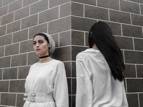 Two individuals stand against a tiled wall, with one facing forward wearing a white textured dress and a headscarf, and the other facing away in a white shirt. The setting conveys a sense of contrast and introspection.