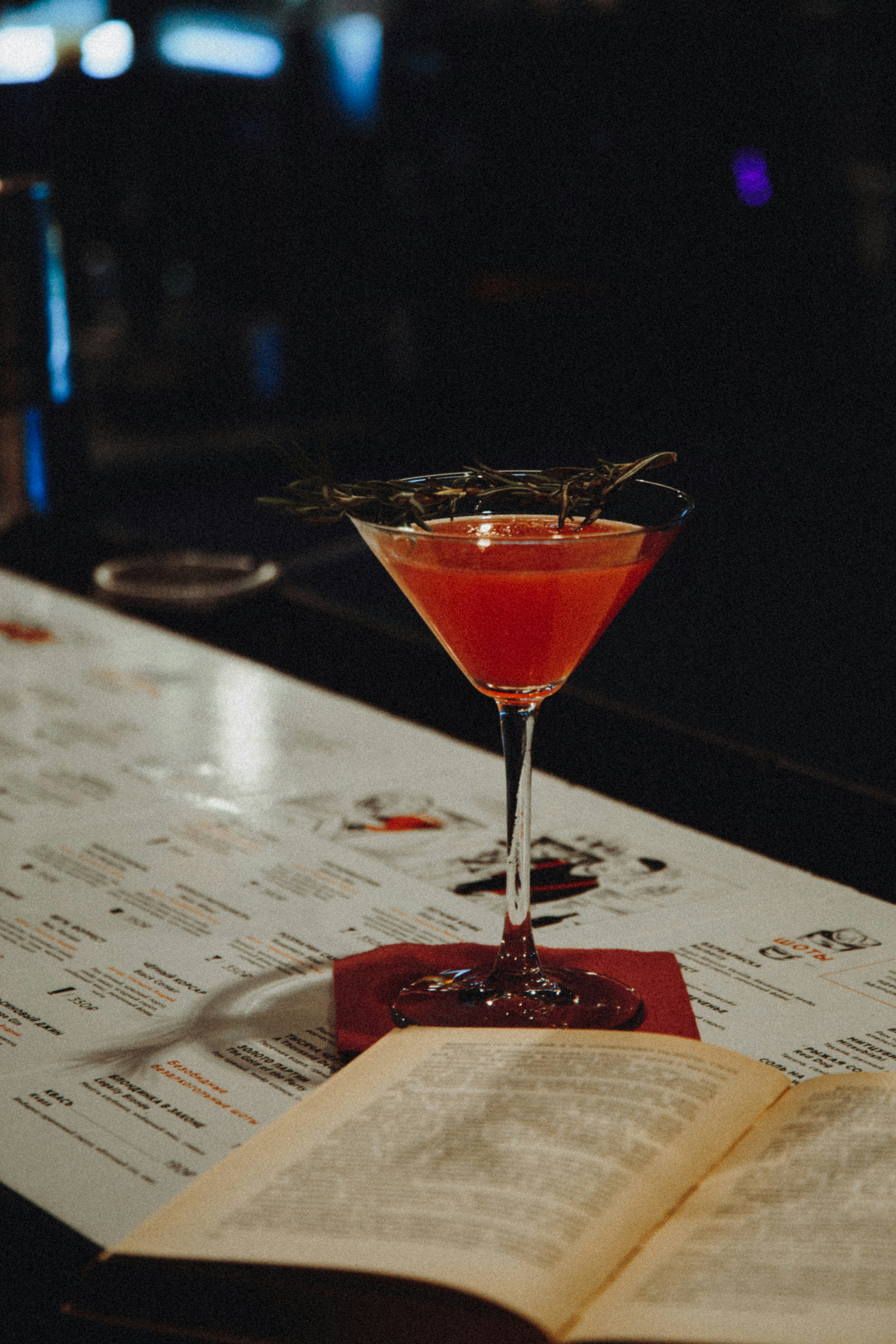 A cocktail glass filled with a vibrant pink drink, garnished with a sprig of herbs, sits on a bar top. An open book lies next to it, with a menu in the background. The setting is dimly lit, creating a cozy and intimate atmosphere.