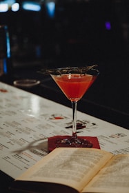 A cocktail glass filled with a vibrant pink drink, garnished with a sprig of herbs, sits on a bar top. An open book lies next to it, with a menu in the background. The setting is dimly lit, creating a cozy and intimate atmosphere.