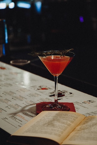 A cocktail glass filled with a vibrant pink drink, garnished with a sprig of herbs, sits on a bar top. An open book lies next to it, with a menu in the background. The setting is dimly lit, creating a cozy and intimate atmosphere.