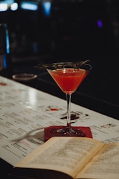 A cocktail glass filled with a vibrant pink drink, garnished with a sprig of herbs, sits on a bar top. An open book lies next to it, with a menu in the background. The setting is dimly lit, creating a cozy and intimate atmosphere.