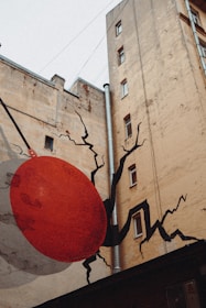 A tall, worn-out brown building with an artistic mural on its wall. The mural depicts a large red ball crashing into the building, creating black cracks that radiate outward. The artwork gives an illusion of structural damage, adding an element of creative urban decoration.