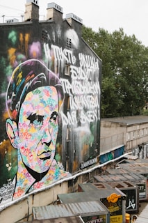 A large, colorful mural on the side of a building features a stylized portrait with vibrant colors. The background includes urban elements like apartment windows and a blend of graffiti and street art. In front of the mural, there are several rooftops of small buildings with signs and urban clutter.