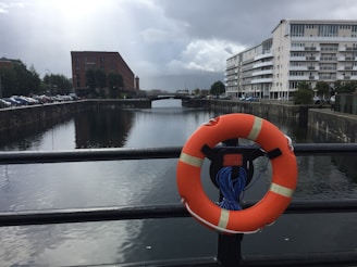 A lifebuoy is attached to a railing overlooking a calm canal flanked by buildings on a cloudy day. A brick building is on the left, and a modern white apartment building is on the right. Cars are parked along the street beside the canal.