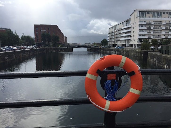 A lifebuoy is attached to a railing overlooking a calm canal flanked by buildings on a cloudy day. A brick building is on the left, and a modern white apartment building is on the right. Cars are parked along the street beside the canal.