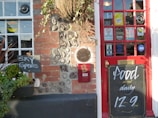 A pub entrance featuring a red door adorned with multiple stickers and signs including 'no smoking' and 'vote now'. Adjacent to the door, there is a chalkboard displaying the message 'Food served daily 12-9 pm'. Beside the entrance, a small blackboard indicates the availability of 'Sky Sports'. The wall consists of red brick and stones with a hanging plant above.