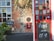 A pub entrance featuring a red door adorned with multiple stickers and signs including 'no smoking' and 'vote now'. Adjacent to the door, there is a chalkboard displaying the message 'Food served daily 12-9 pm'. Beside the entrance, a small blackboard indicates the availability of 'Sky Sports'. The wall consists of red brick and stones with a hanging plant above.